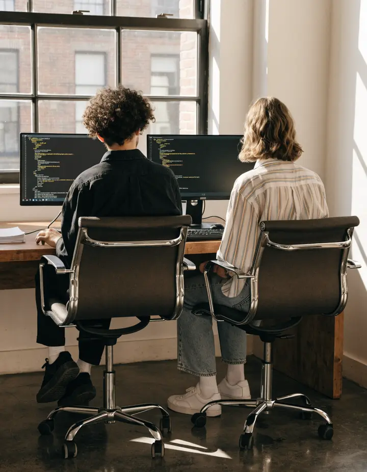 Two people pair programming at a window desk, seen from behind