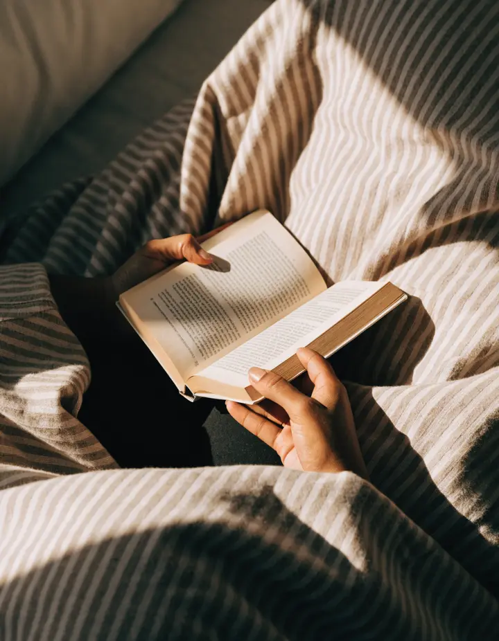 A reader under a striped blanket holding an open paperback