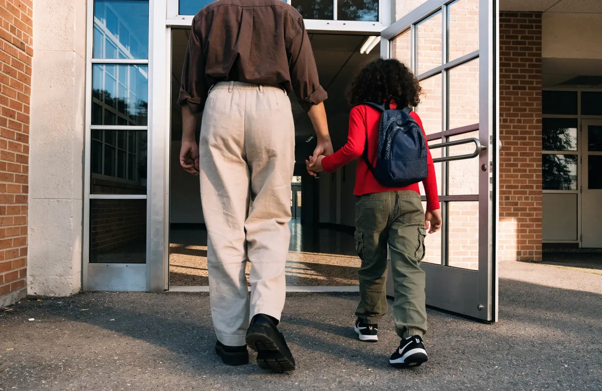 A parent and child walking hand in hand into a brick school building, seen from behind