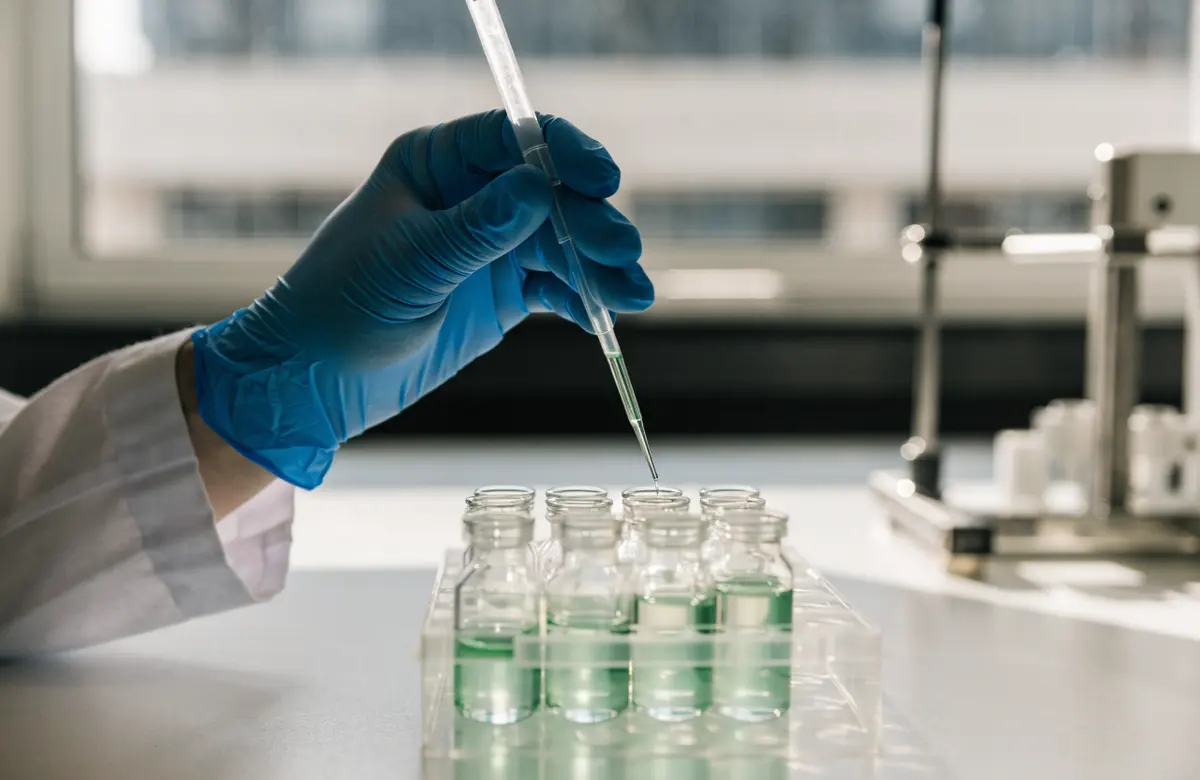 Gloved hands pipetting a green sample into vials at the testing lab